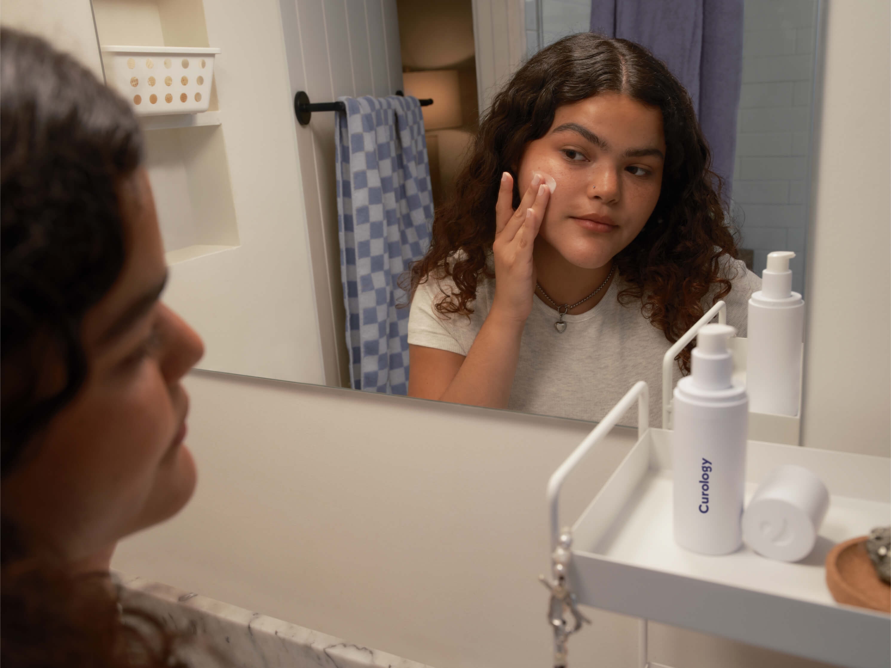 Young brunette woman with curly hair, looking in the bathroom mirror while applying Curology Custom Formula, with the uncapped open bottle on a white shelf next to her.