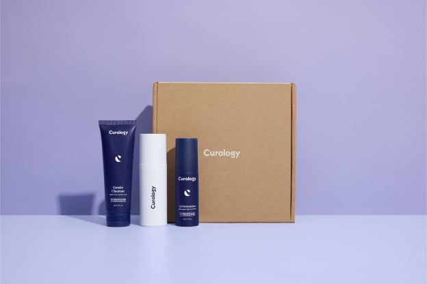 Purple Curology Cleanser tube, white Curology Custom Formula Rx bottle, and purple Curology Moisturizer bottle standing to the right of a square brown Curology packaging box on a light purple background.